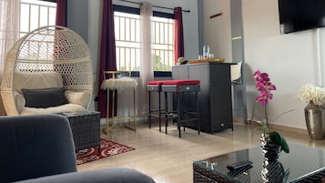 Una sala de estar moderna con un sofá de mimbre, una mesa de centro de vidrio, una zona de bar con taburetes y un televisor montado en la pared.