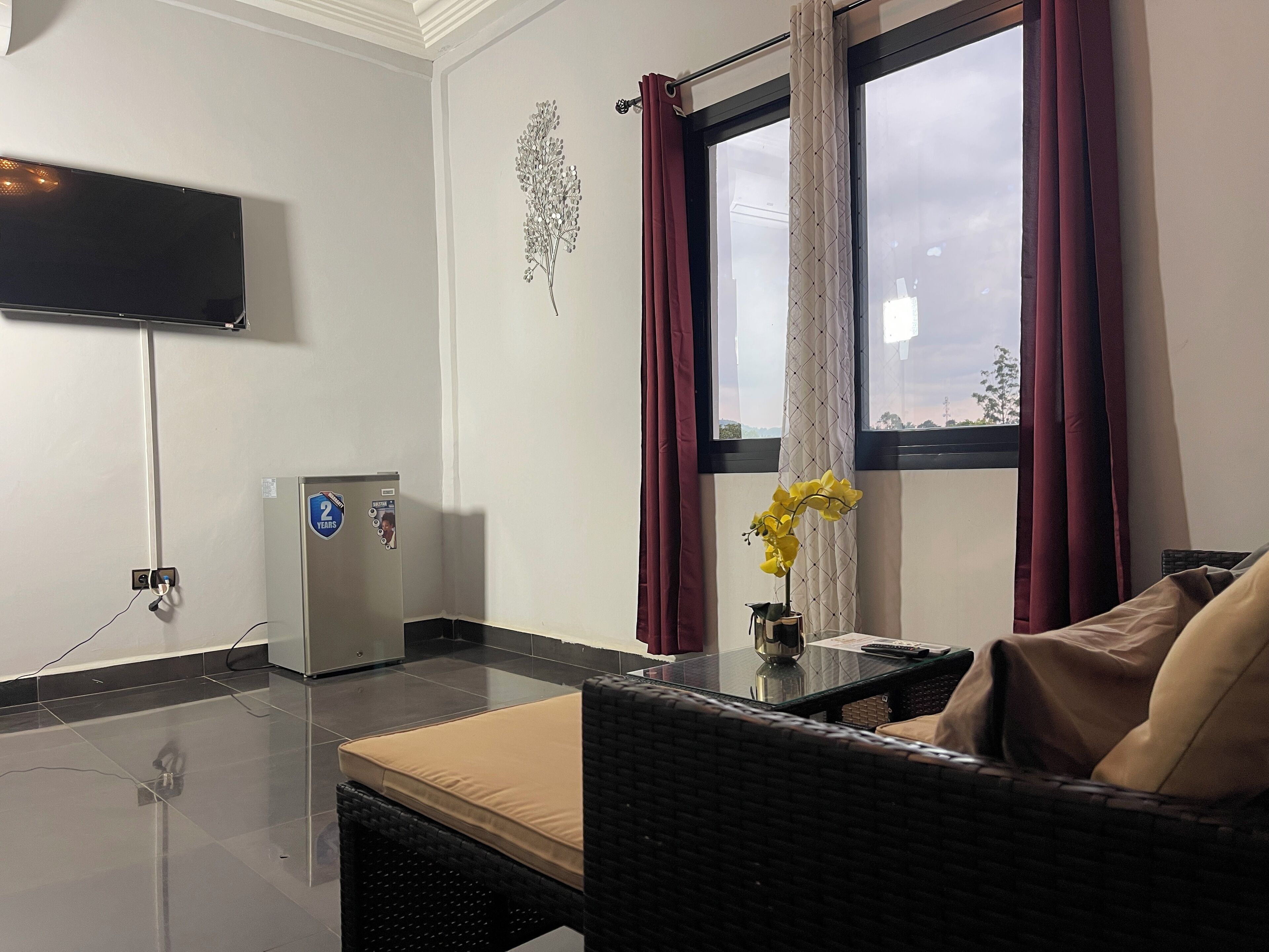 Une chambre d’hôtel équipée d’un téléviseur à écran plat fixé au mur, d’un petit réfrigérateur, d’une table en verre ornée d’un vase de fleurs jaunes et d’un canapé en osier avec un coussin beige.
