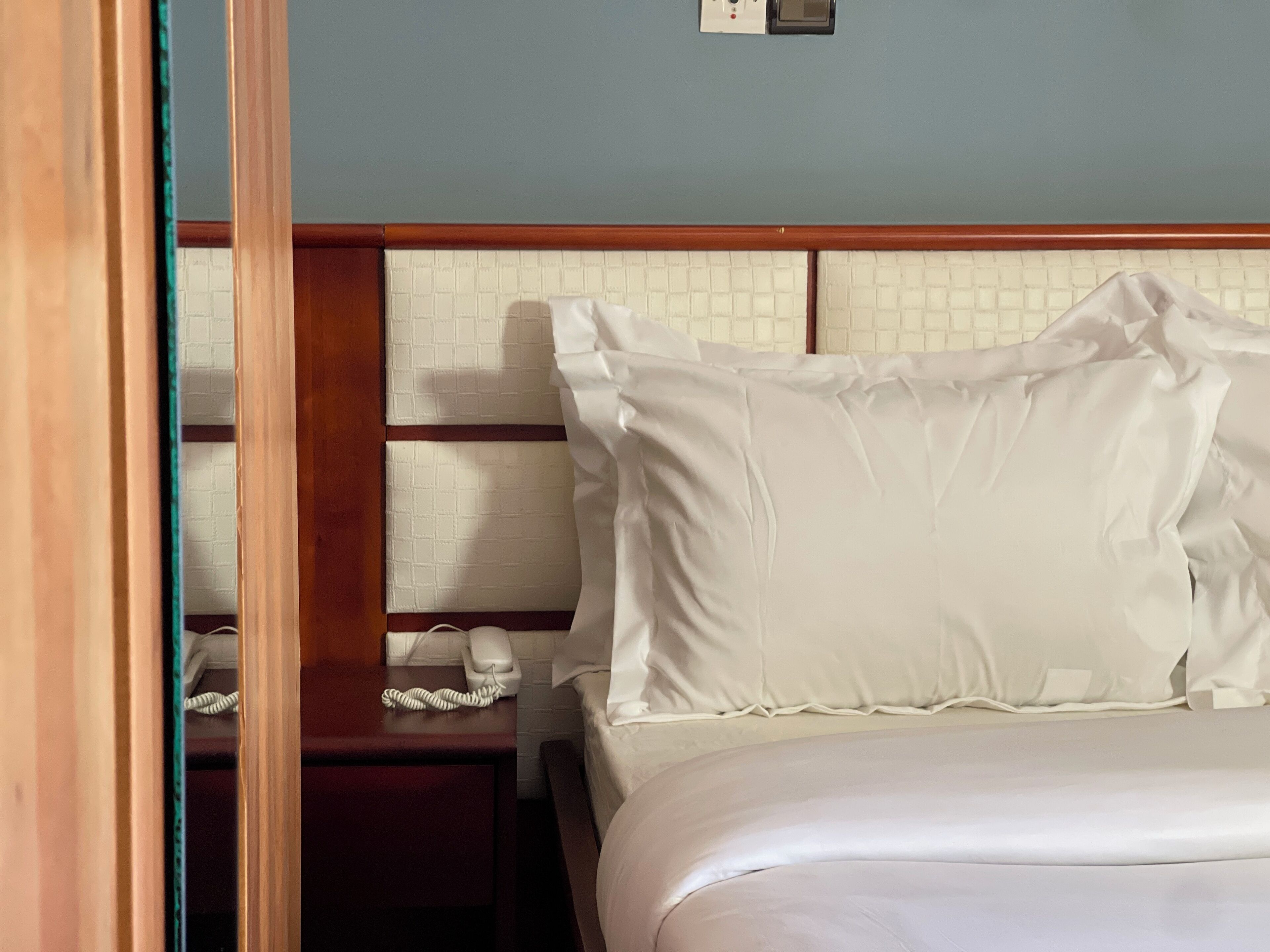 A hotel room with a bed, a wooden headboard, a nightstand with a telephone, and a wall-mounted light switch.