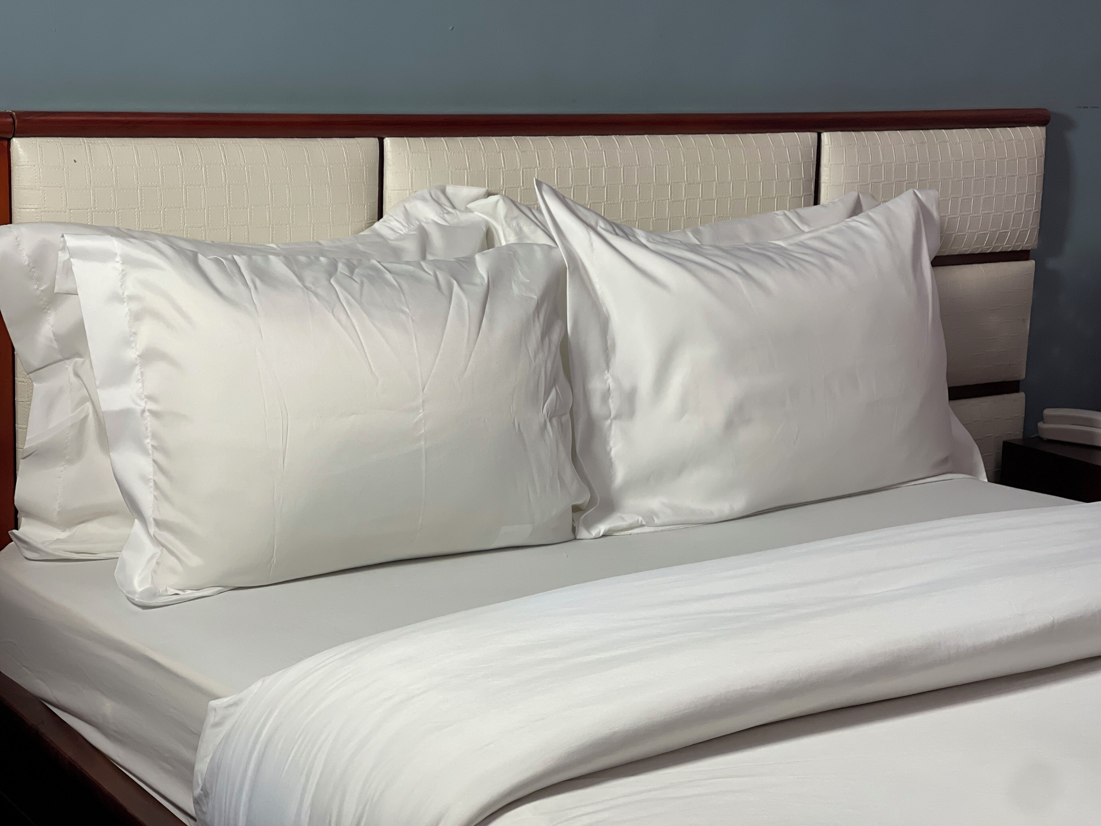Uma cama bem arrumada com lençóis e almofadas brancos, uma cabeceira de madeira e uma mesa de cabeceira com um telefone.