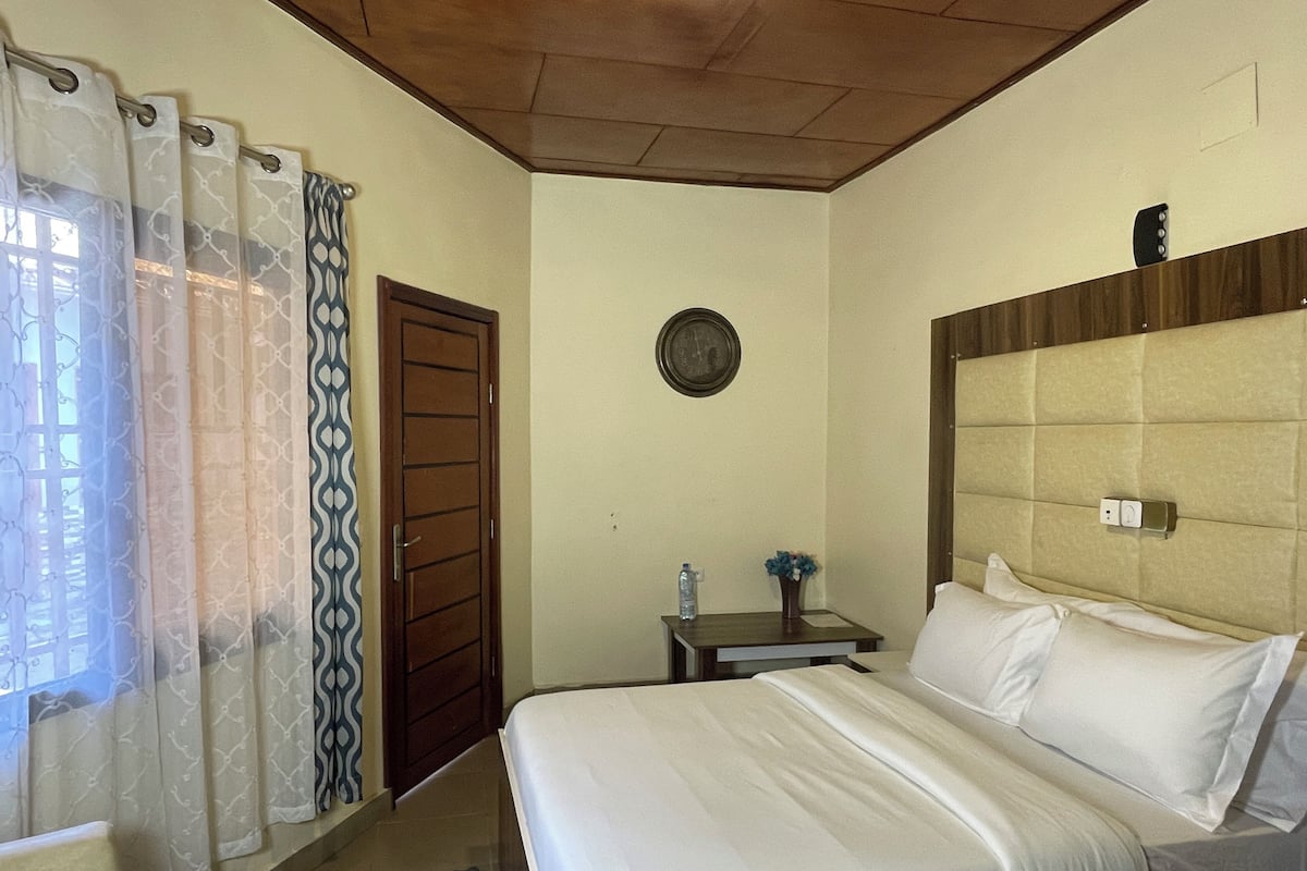 Habitación de hotel con cama, mesita de noche, reloj de pared y ventana con cortinas estampadas.