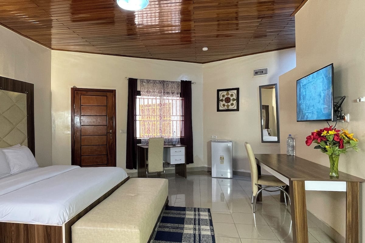 Habitación de hotel con cama, escritorio, silla, refrigerador pequeño, espejo y ventana con cortinas.