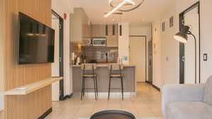 A modern living room with a flat-screen TV, a grey sofa, and a kitchen area with a microwave and bar stools. - Incredible 2BR With Balcony in San Isidro (Lima)