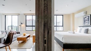 Quarto de hotel moderno com uma cama grande, um sofá, uma mesa de jantar e vista da cidade através de amplas janelas.
