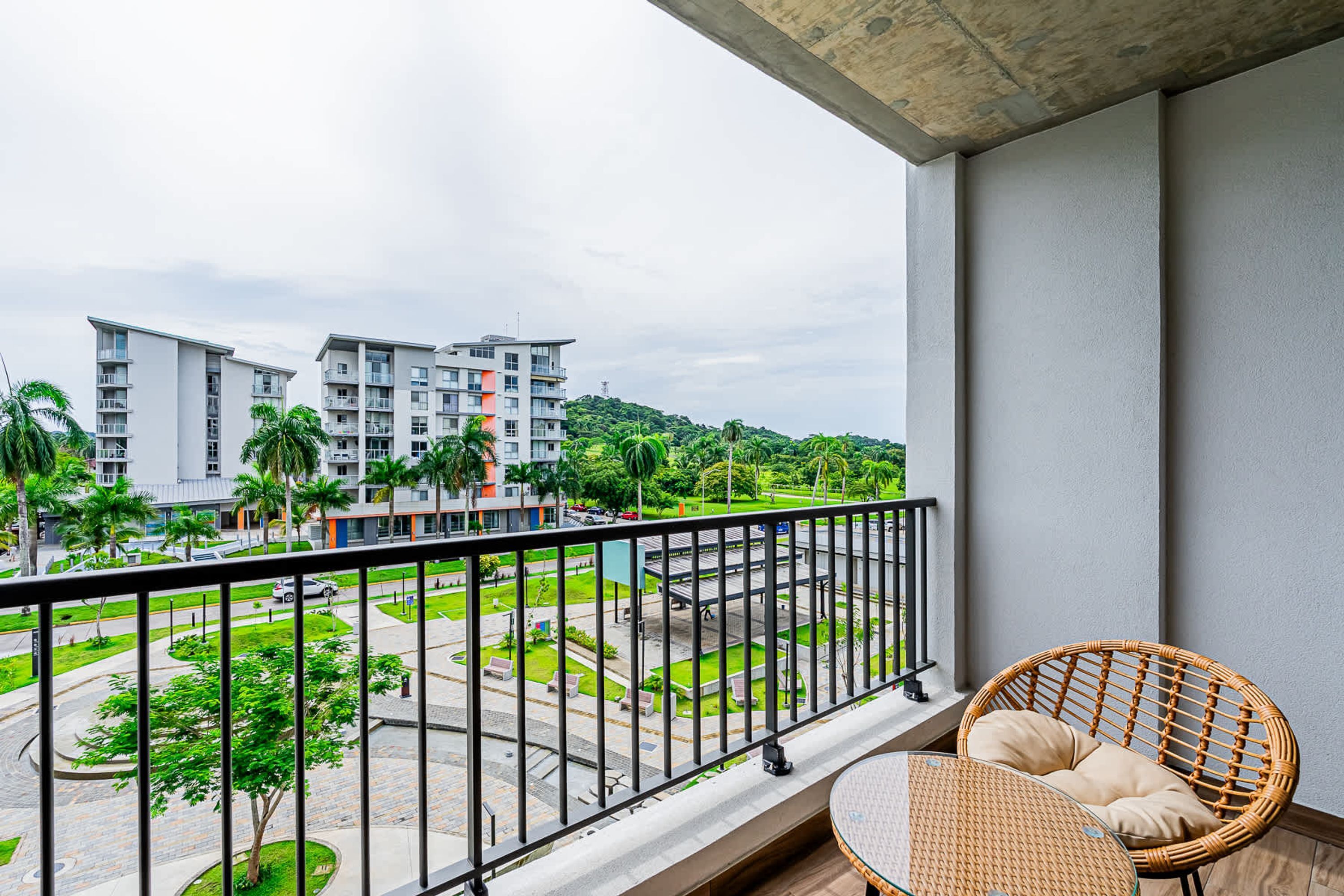 A balcony with a wicker chair, a view of a landscaped area, and modern buildings in the background.