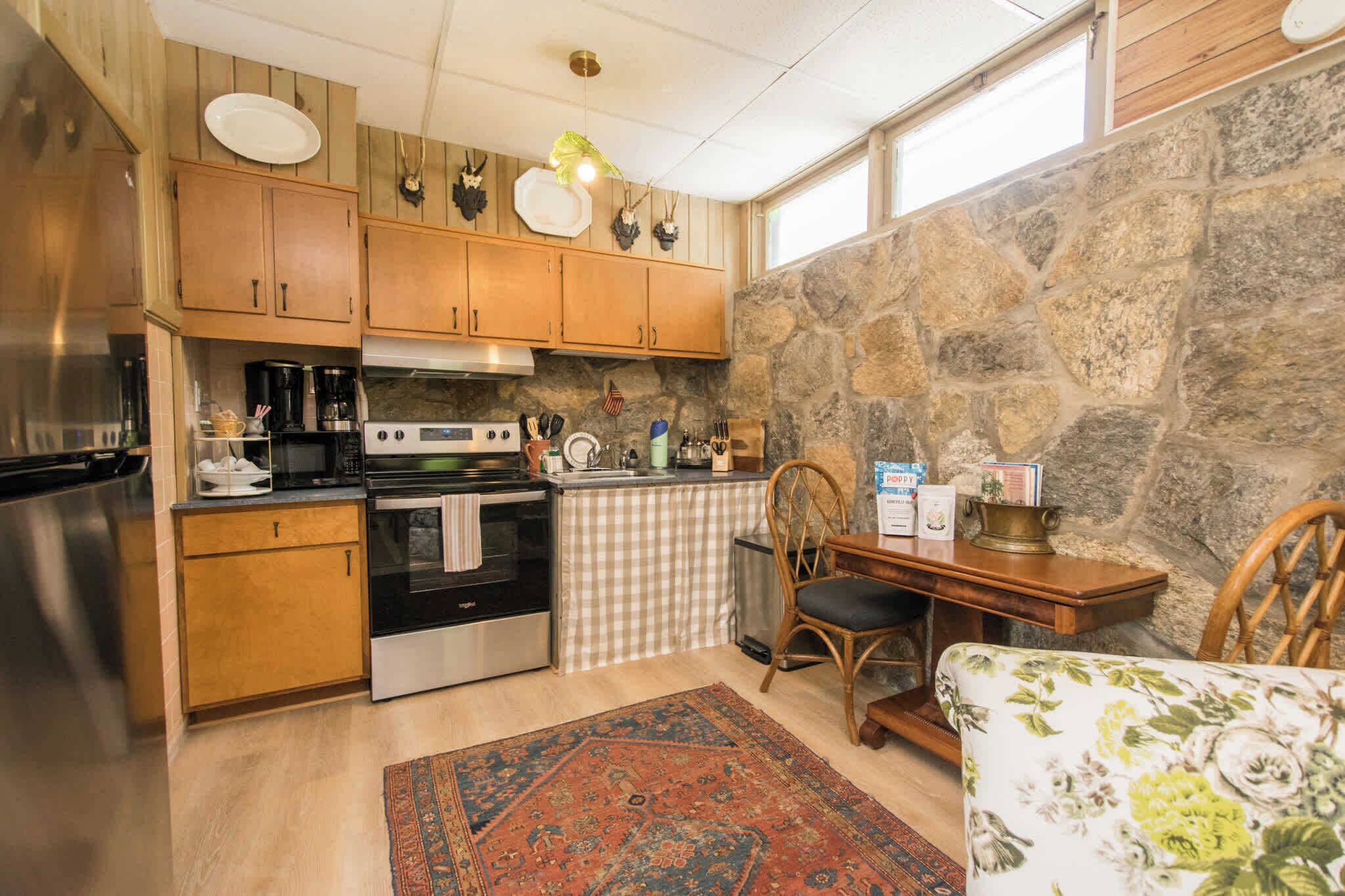A compact kitchen with wooden cabinets, a stone wall, a checkered tablecloth, and a floral-patterned chair.