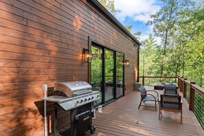 Una terraza de madera con una parrilla, asientos exteriores y un ventanal que da a otra zona de terraza.