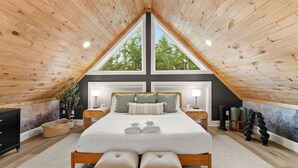 A bedroom with a sloped ceiling, a large bed with pillows, bedside lamps, and a view through a window.