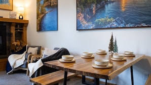Un coin repas avec une table en bois, dressée avec des bols et des assiettes, une cheminée et deux grandes peintures représentant des paysages accrochées au mur.