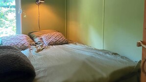 Un lit simple avec une couette blanche et deux coussins à motifs, une lampe de chevet et une vue sur la verdure à l’extérieur.
