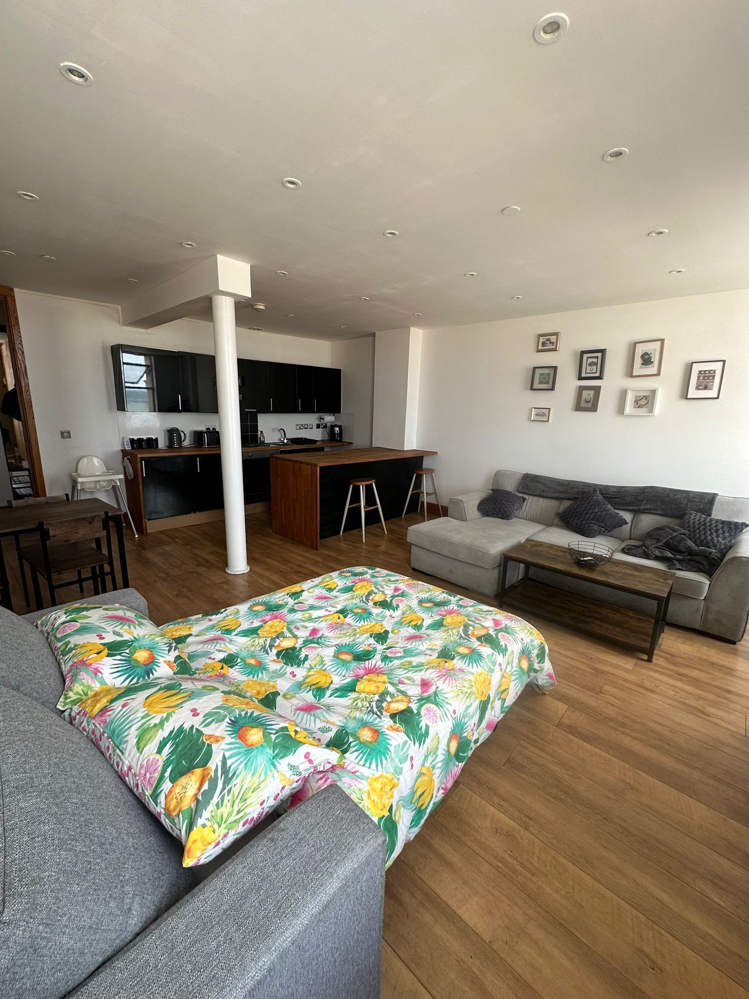 Ein geräumiger Wohnbereich mit einer Couch, einer farbenfrohen blumigen Tagesdecke, einem Holztisch und einem grauen Sofa. Im Hintergrund ist eine Küche mit dunklen Schränken und einer weißen Arbeitsplatte zu sehen.