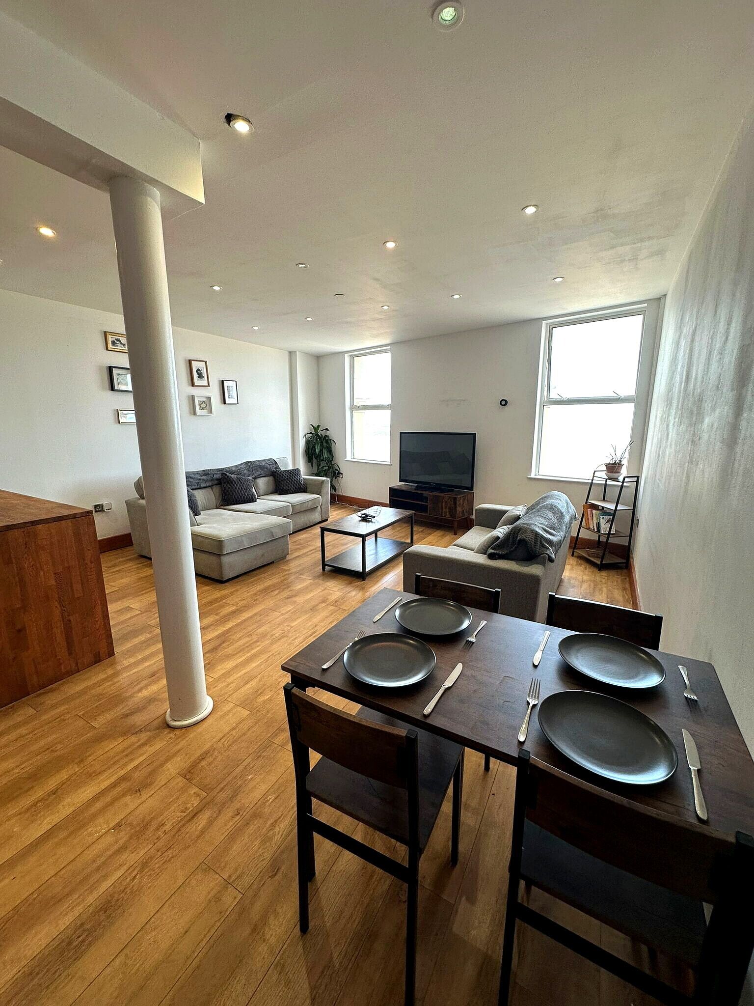 A modern living room with a dining area, wooden flooring, and a large window providing natural light.