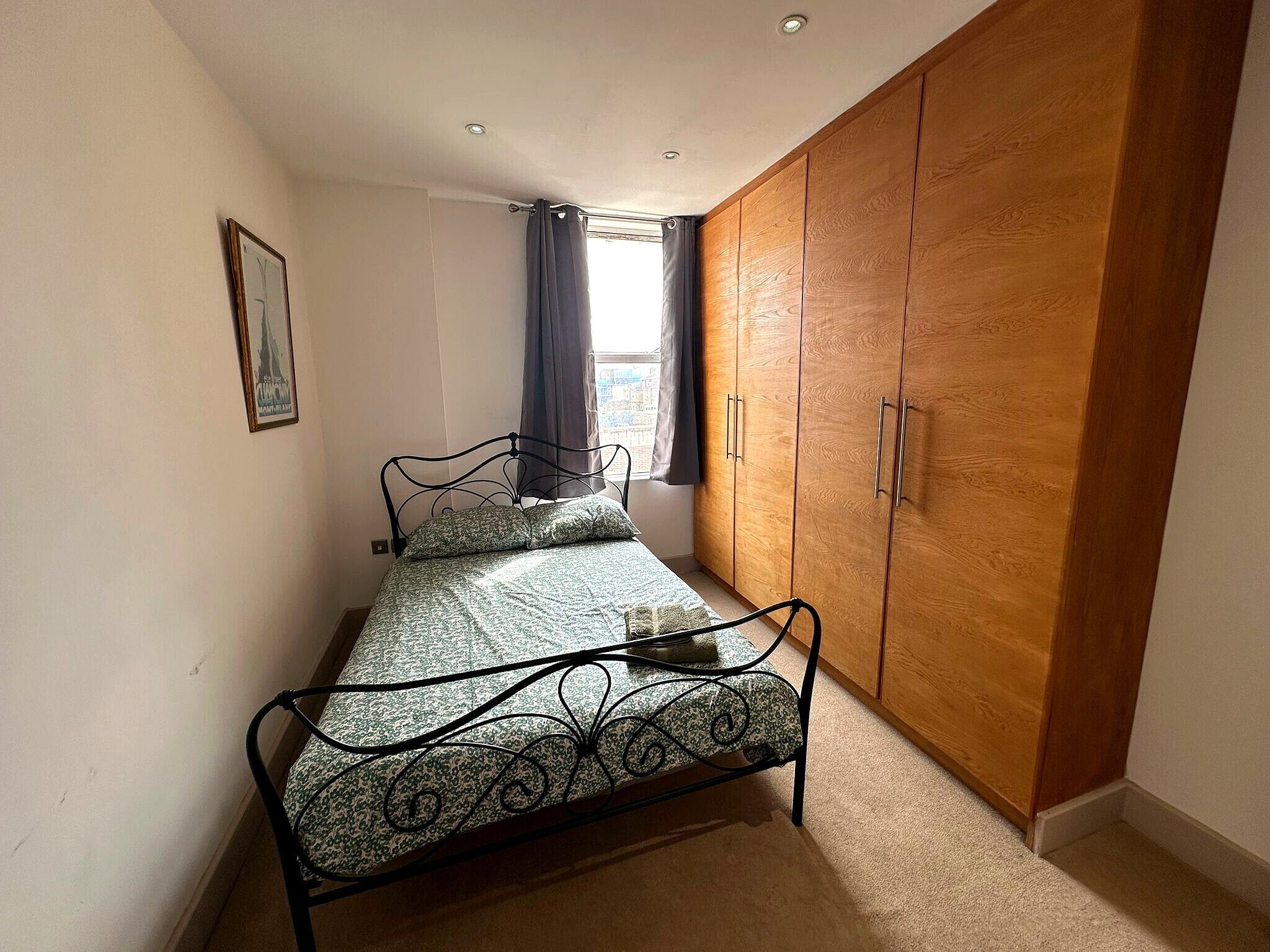 Une petite chambre à coucher avec un lit en métal, une armoire en bois et un seul tableau encadré accroché au mur.