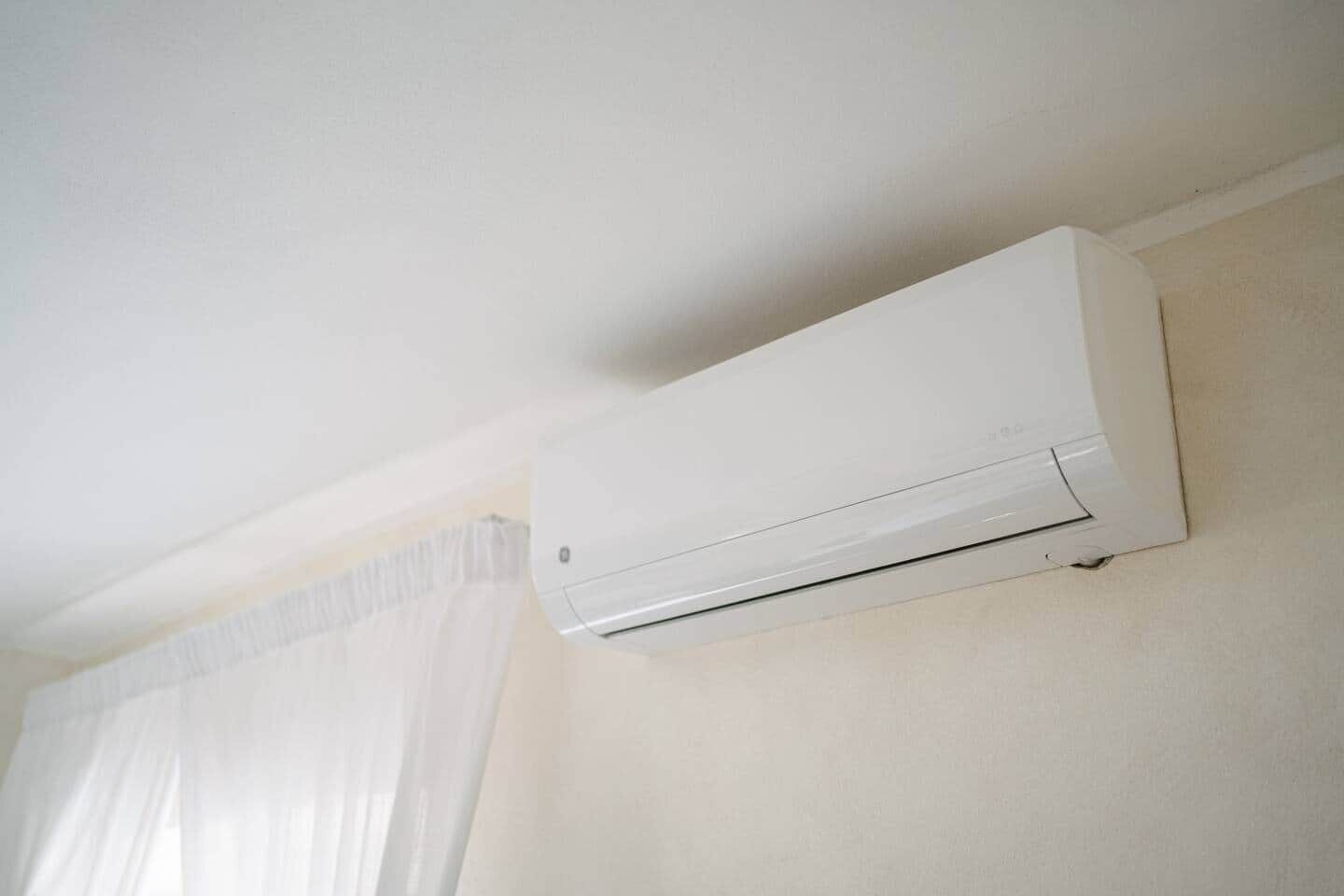 A white air conditioning unit mounted on a wall in a room with a window and curtains.