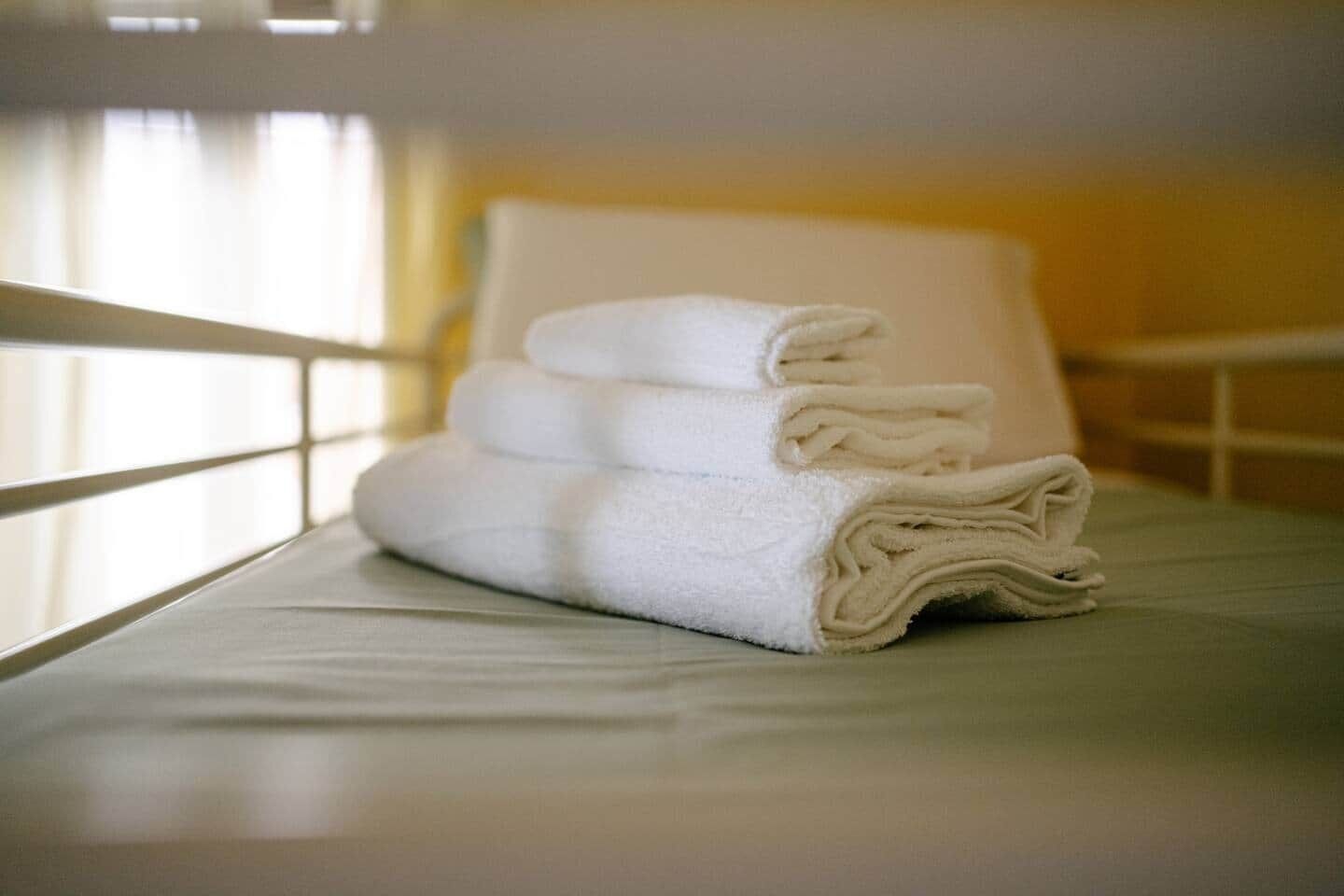 Uma pilha organizada de toalhas brancas sobre uma cama com cabeceira branca e uma janela com cortinas.