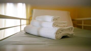 Uma pilha organizada de toalhas brancas sobre uma cama com cabeceira branca e uma janela com cortinas.