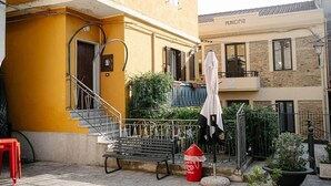 Un edificio amarillo con una escalera, un banco, un bote de basura rojo y una zona de estar al aire libre techada.