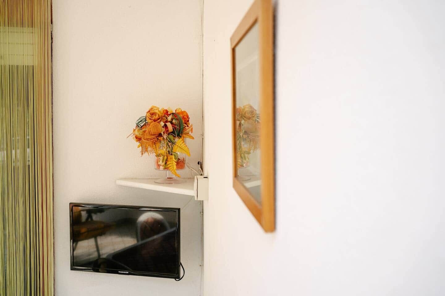 A white wall with a flat-screen TV mounted below a shelf, a framed mirror, and a vase with flowers on the shelf.