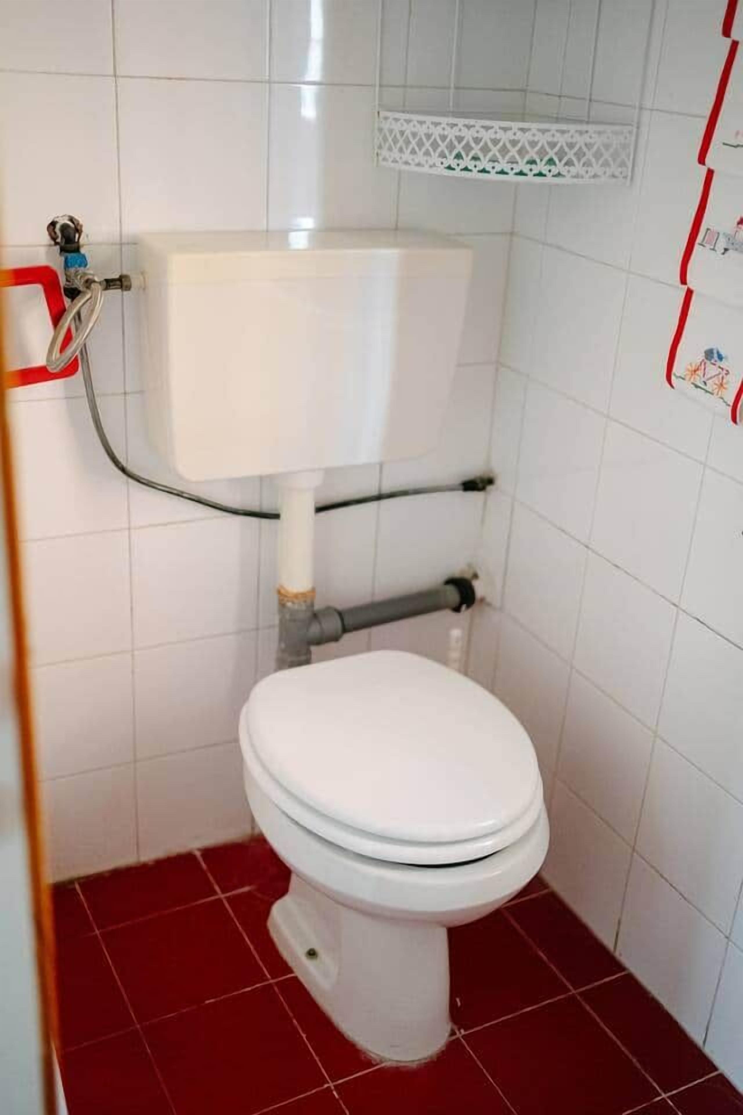 Un baño pequeño con inodoro blanco, baldosas de suelo rojas y pared revestida de azulejos blancos.
