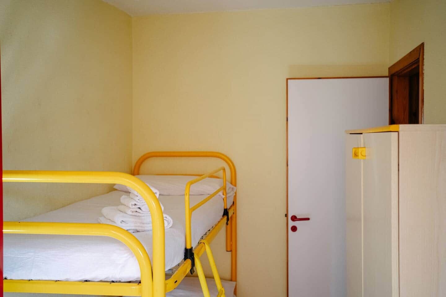 Una cama individual con estructura de metal amarillo, una puerta blanca con picaporte rojo y un armario blanco.