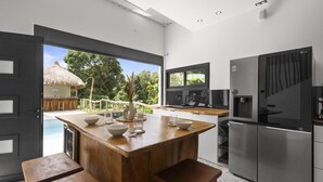Une cuisine moderne dotée d’un îlot central en bois, d’appareils électroménagers en acier inoxydable et offrant une vue sur une piscine et une structure à toit de chaume.