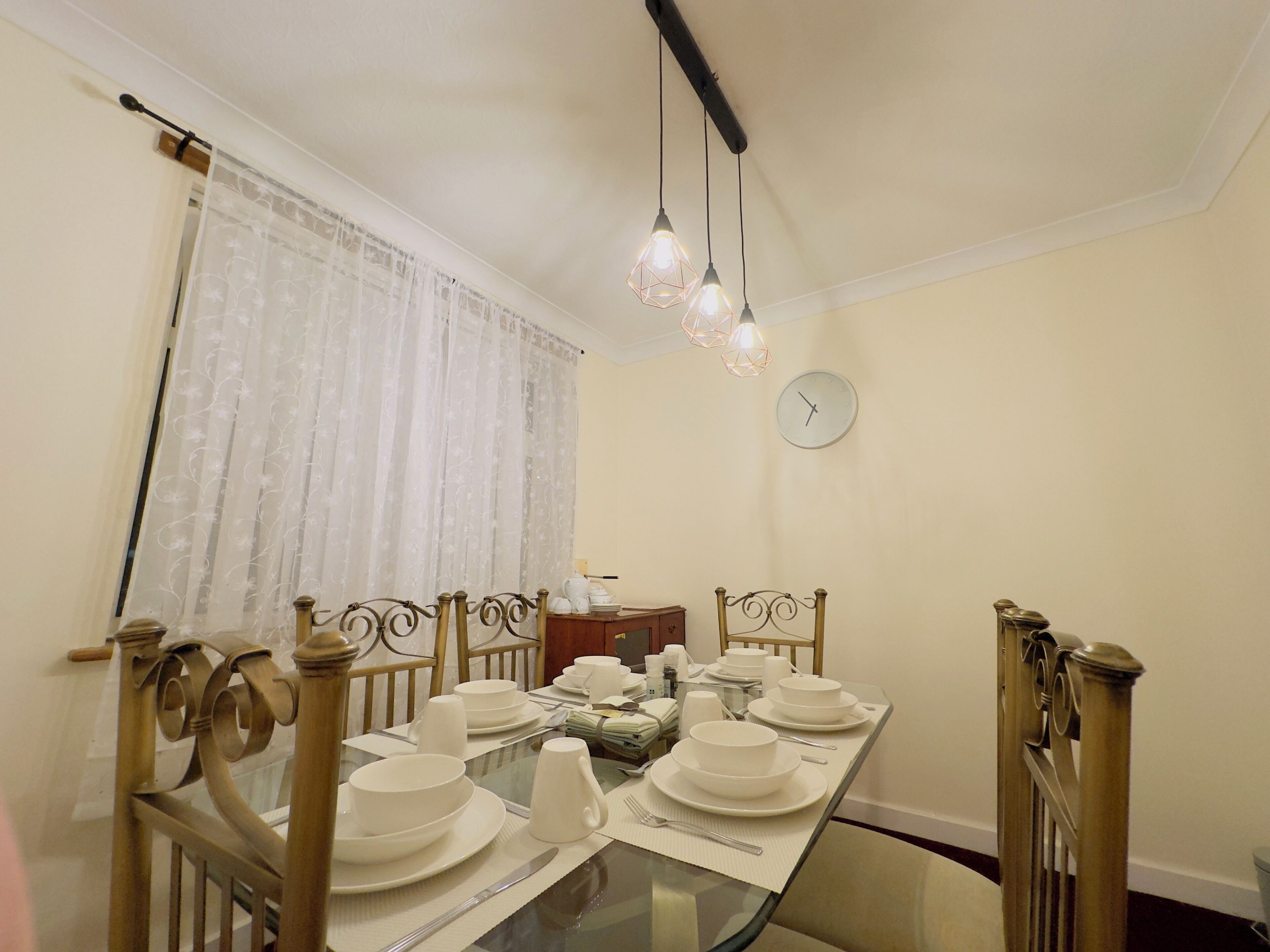 A dining area with a glass table set for four, surrounded by wooden chairs with intricate designs. A clock is mounted on the wall above the table.