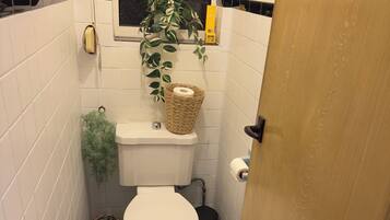 A small bathroom with white tiled walls, a white toilet, a black trash bin, a wicker basket with toilet paper, a small green plant, and a wooden door.