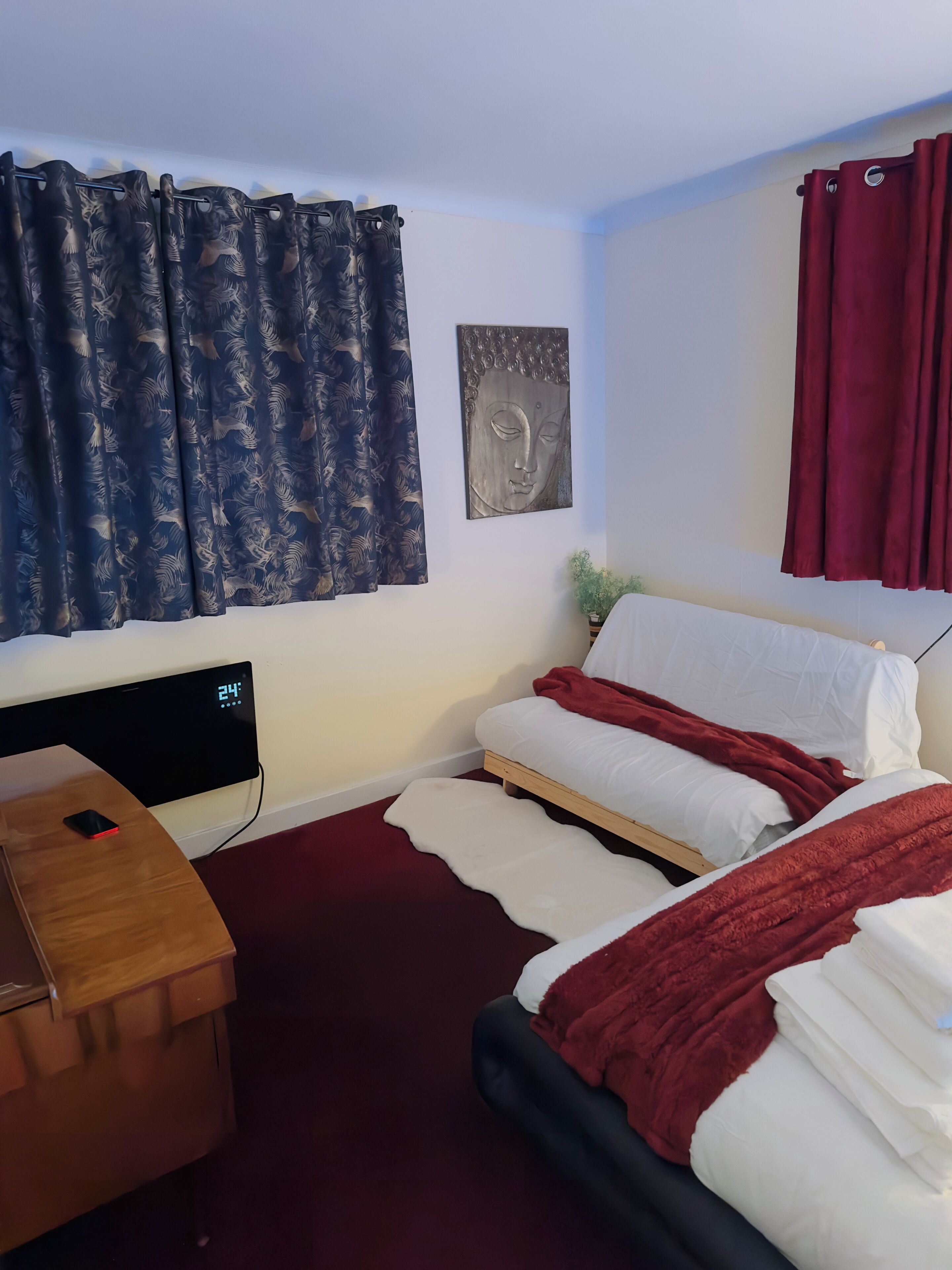 A bedroom with a bed, a bedside table, a wall clock, and a window with curtains.