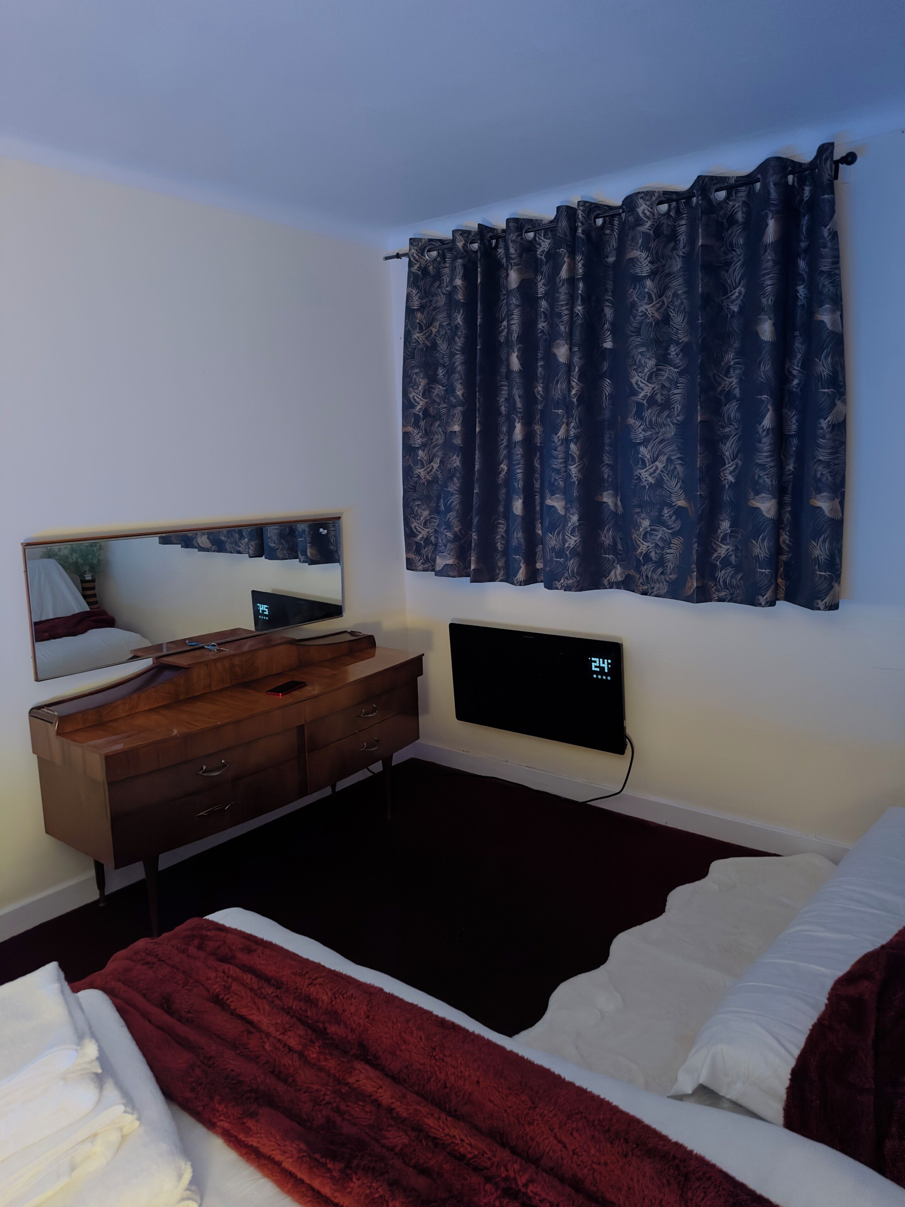 A bedroom with a bed, a bedside table, a mirror, a TV, and a window with curtains.