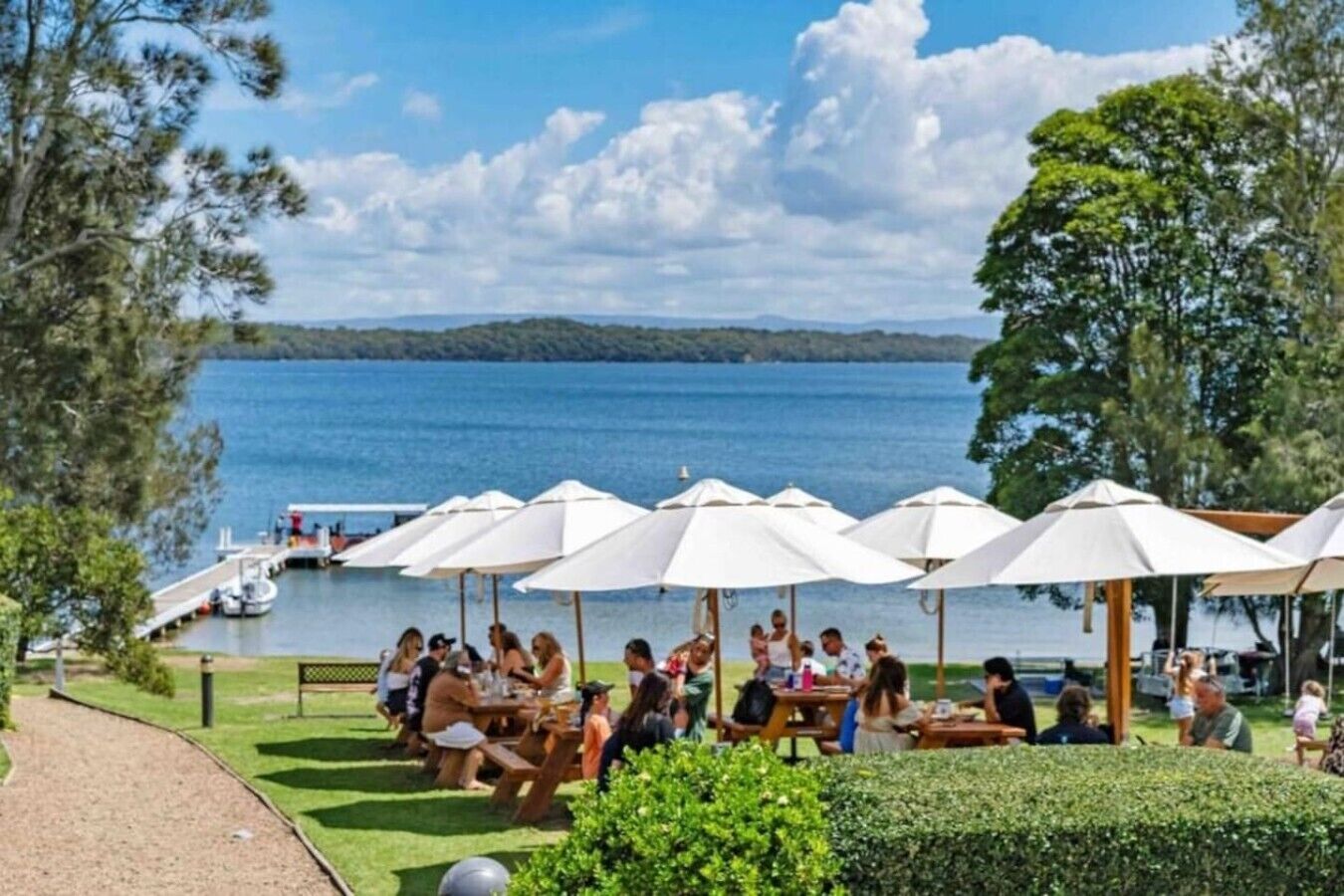 Un espace de restauration au bord du lac, avec des tables à l’extérieur et des parasols, un quai avec des bateaux, et une vue sur le lac et les collines lointaines.