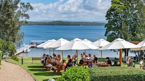 A lakeside dining area with outdoor seating and umbrellas, a dock with boats, and a view of the lake and distant hills. - Elysian Escape  - 4 Pools, Lakeside, Raffertys Res (Cams Wharf)