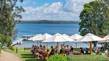 Un espace de restauration au bord du lac, avec des tables à l’extérieur et des parasols, un quai avec des bateaux, et une vue sur le lac et les collines lointaines.