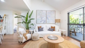 A modern living room with a white sofa, a round wooden coffee table, a large plant, and a balcony with outdoor furniture. - Elysian Escape  - 4 Pools, Lakeside, Raffertys Res (Cams Wharf)