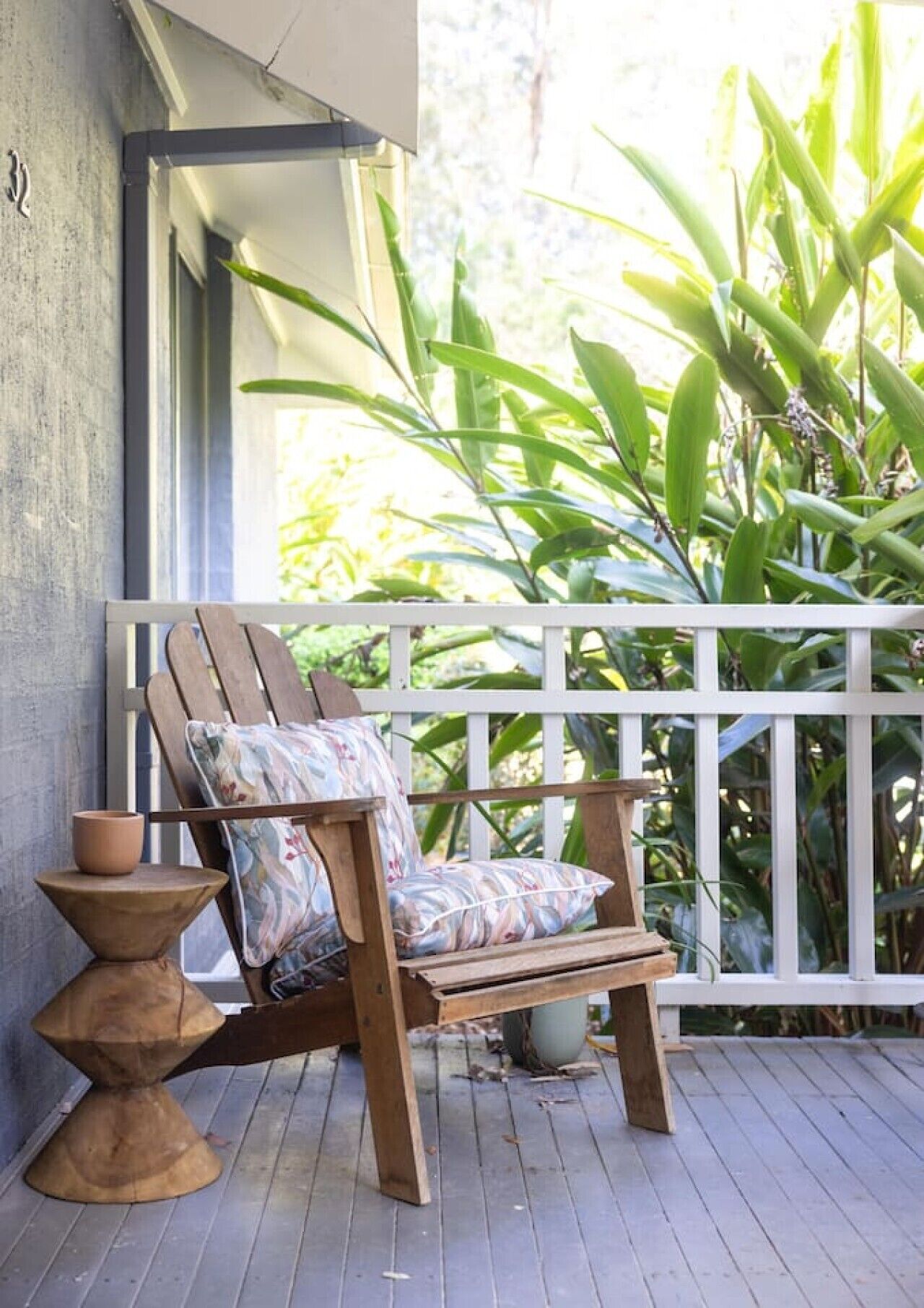 A wooden chair with a floral cushion on a wooden deck, a small round table with a terracotta pot, and a white railing with greenery in the background.