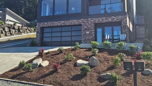 A modern two-story house with a large garage, stone facade, and a well-maintained garden with rocks and shrubs. - Industrial modern house with Water Views – Walk to the waterfront (Tacoma)