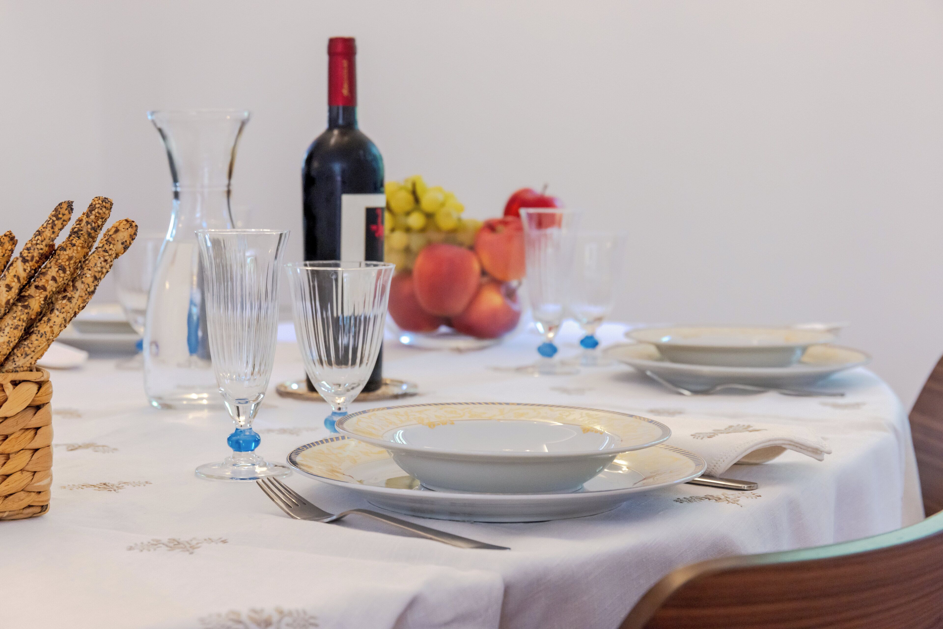 A dining table set with plates, cutlery, glasses, and a bottle of wine, with fruits and bread in the background.