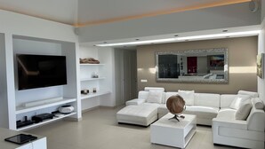A modern living room with a flat-screen TV, white sofas, a coffee table, and a large mirror.