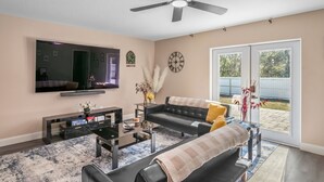 Ein Wohnzimmer mit Flachbildfernseher, Deckenventilator, Sofa, Couchtisch und einem großen Fenster mit Blick ins Freie.