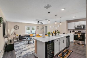 A modern kitchen with white cabinets, a built-in oven, and a dishwasher. There is a dining area with a black sofa and a coffee table. - 4 BR Family Home , EV Charger, Kids & WFH. (Ocala)