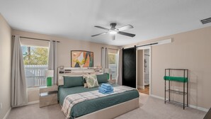 Ein Schlafzimmer mit Bett, Deckenventilator, Fenster mit Vorhängen, Nachttisch, Wandregal und einer Tür zu einem anderen Raum.