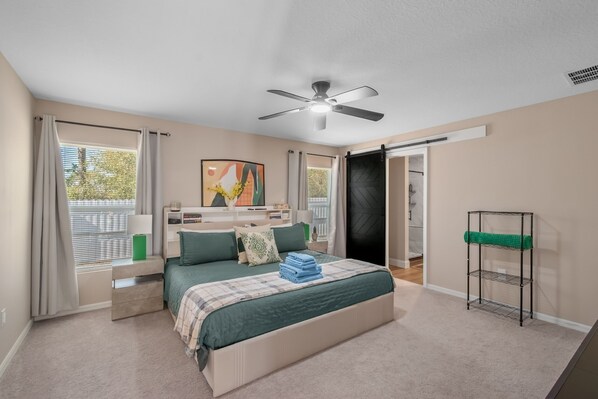 Une chambre à coucher comprenant un lit, un ventilateur de plafond, une fenêtre avec des rideaux, une table de chevet, une commode, une étagère fixée au mur et une porte donnant sur une autre pièce.