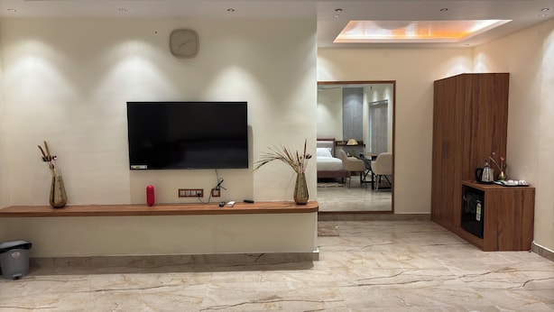 Una sala moderna con un televisor de pantalla plana, un mueble bajo de madera, un jarrón con plantas secas y un espejo que refleja un dormitorio con una cama y una mesa.