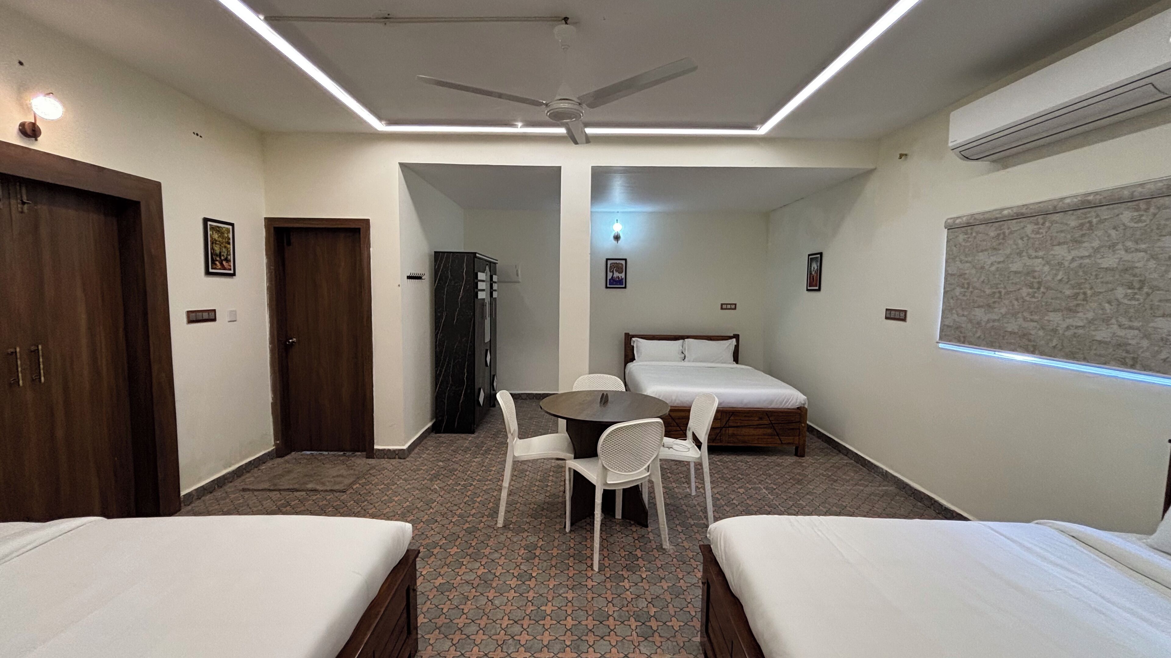 A hotel room with two single beds, a small table with chairs, a ceiling fan, and a window with blinds.