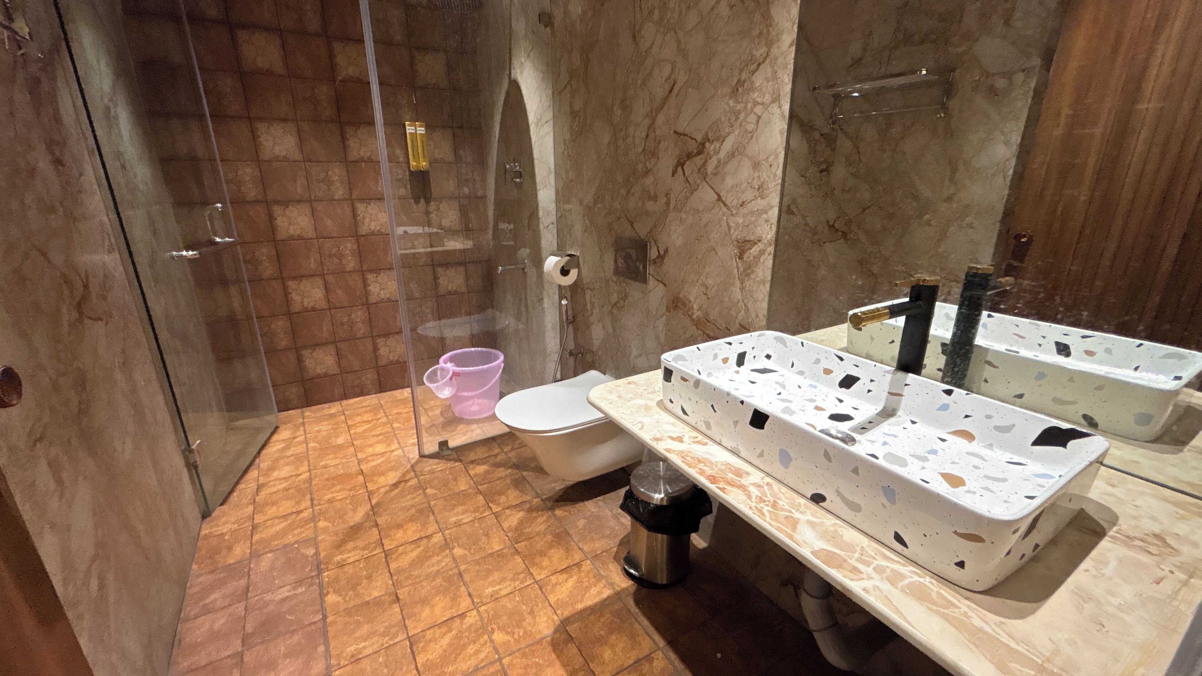 A bathroom with a glass shower enclosure, a toilet, a sink with a marble countertop, and a trash can.
