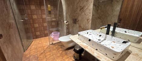A bathroom with a glass shower enclosure, a toilet, a sink with a marble countertop, and a trash can.