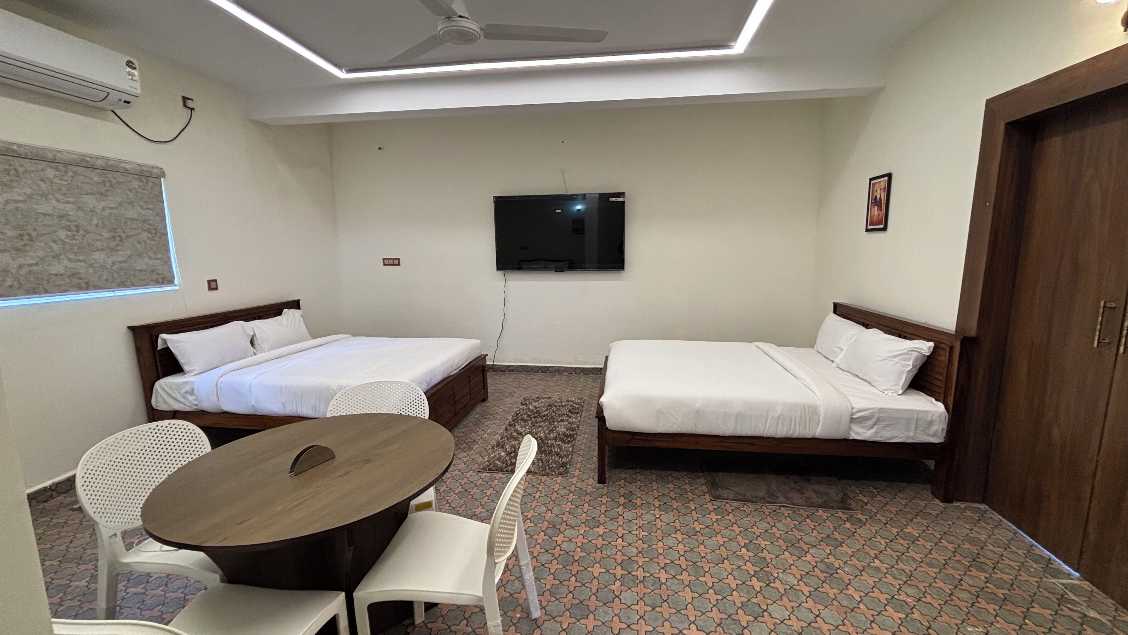 A compact hotel room with two single beds, a small table, two chairs, a ceiling fan, a TV, and a wardrobe.