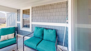 A covered porch with a green sofa and a chair, a window, and a wall with gray shingles. - KH3111B: Seas the Day | Walk to Beach | Patio | Kitchenette (Kitty Hawk)