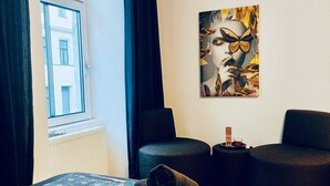 Habitación de hotel con cama, ventana, un cuadro, dos sillas y una mesita con una vela.