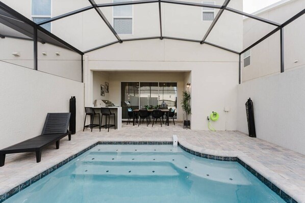 Une piscine avec un patio couvert, une table à manger avec des chaises et un fauteuil de relaxation.