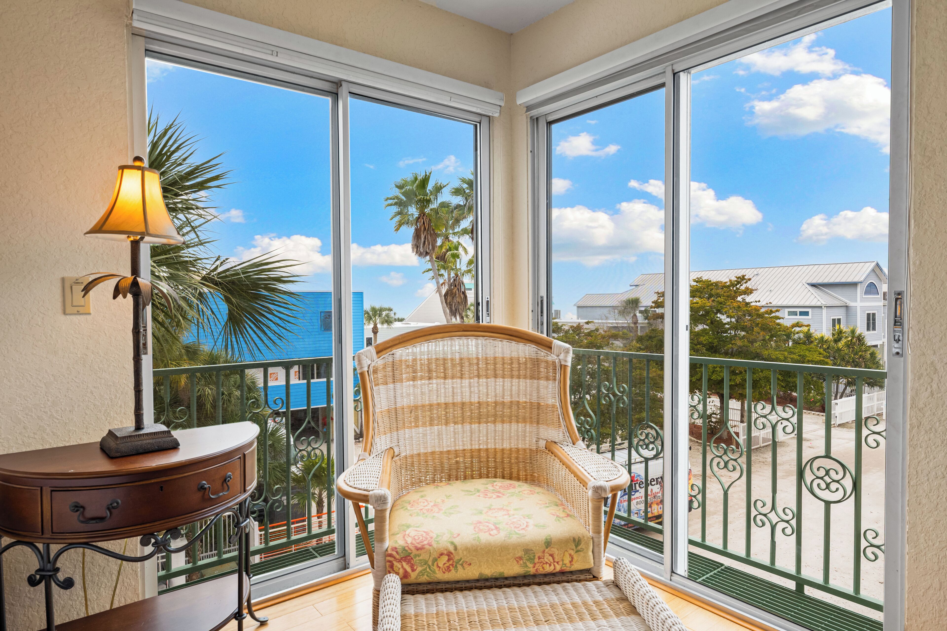 A balcony with a wicker chair, a small table with a lamp, and a view of the ocean and buildings.